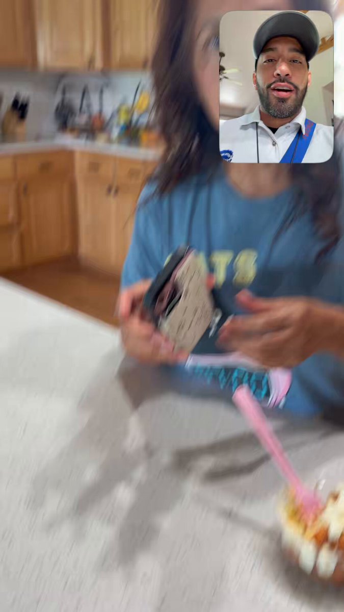 The image shows a man talking into a video call with a woman, who appears to be a young girl wearing a blue shirt. The man has a camera in his hand, possibly showing off something interesting or entertaining for the woman. In the background, there is a dining table and various kitchen items such as knives and spoons. A cake can also be seen on the table, suggesting that they might have been celebrating a special occasion like a birthday.