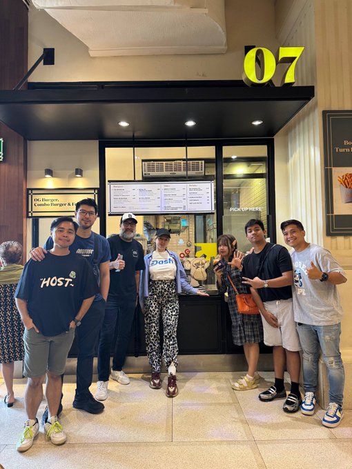 A group of seven people pose cheerfully in front of "07 Bored & Hungry Burger" restaurant, with a woman in a "Dash" shirt making a peace sign at the center, alongside a visible menu board and "PICK UP HERE" sign. The image ties to the post by capturing the team's gathering at the Web3-themed burger joint in Manila to discuss crypto merchant adoption challenges.