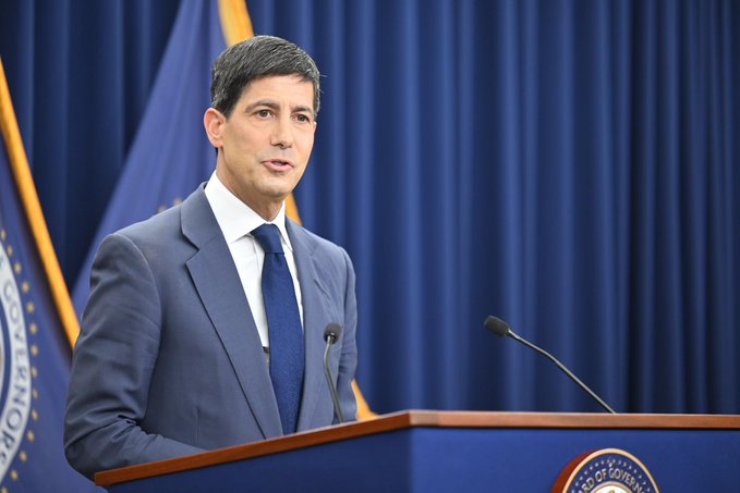 A man in a blue suit stands at a podium speaking, with a blue curtain background and flags, including what appears to be a Federal Reserve seal.