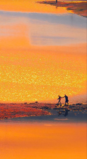 Two silhouetted figures appear to be holding hands while running across a beach at sunset, with the orange water reflecting the golden light in a shimmering pattern.