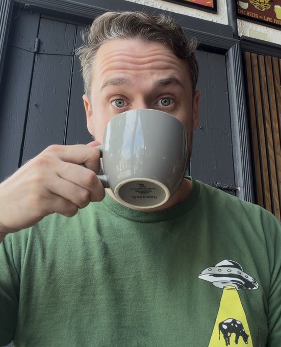 A man drinking from a grey 'THING&CUP' mug while wearing a green sweatshirt featuring a UFO abducting a cow graphic, sitting in what appears to be a pub.