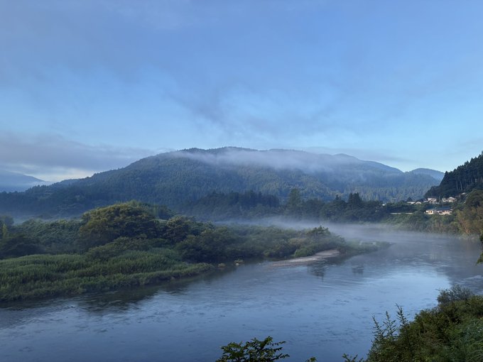 A serene landscape view of a river flowing through a valley with mist-covered green mountains and forested hills under a partly cloudy blue sky.