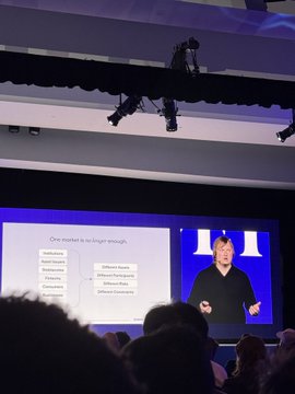 The image shows a man giving a presentation with a large screen behind him displaying his slides, which include information about building onchain credit markets for internet scale. The room appears to be filled with people who are listening attentively to the speaker.