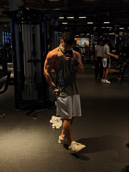 A muscular man takes a mirror selfie in a dimly lit gym, flexing his arms while wearing a green "MEN'S HEALTH" tank top and gray shorts, with workout equipment and other gym-goers visible in the background—capturing the start of a "GM" (Good Morning) fitness routine.