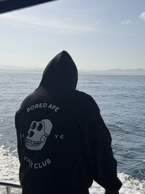 A person wearing a black "Bored Ape Yacht Club" (BAYC) hoodie stands on a boat, gazing out at the open ocean under a partly cloudy sky, embodying the post's claim about repping BAYC both online and IRL before a great white shark cage dive.