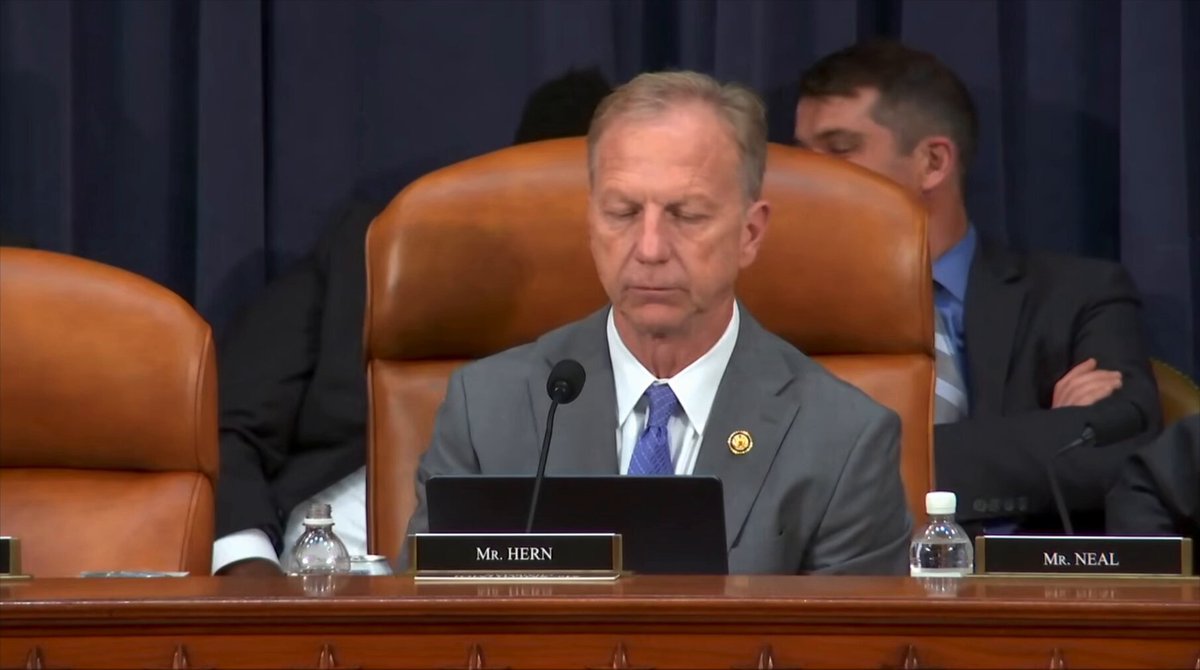 The image shows a man sitting at a desk, wearing a suit and tie, with his hands clasped together as he appears deep in thought or concentration. The setting seems to be an office environment, possibly a conference room where people are gathered for a meeting or discussion. There is another person visible in the background, but they are not the main focus of the image.