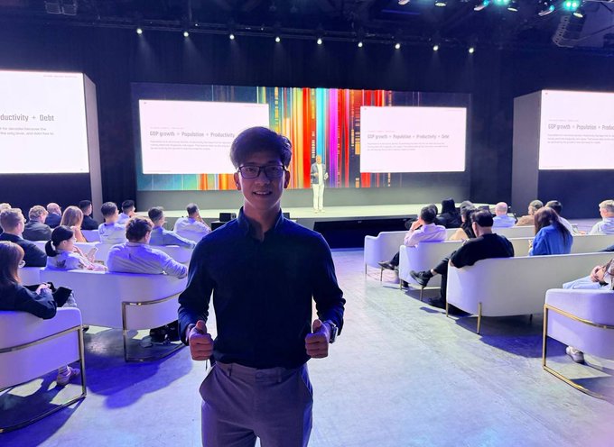 A young man wearing glasses and a dark shirt gives two thumbs up while posing in front of a conference stage with screens displaying "GDP growth = Production = Productivity = Unit."