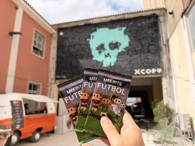 The image shows a person holding three bags of Meebits Fútbol, which are soccer-themed chocolate bars. The bags have the text "MEEBITS FU TOL" printed on them. The person is standing outside in front of a building with a mural featuring a skull and crossbones design. The post also mentions that there will be a free giveaway of these Meebits Fútbol packs at an event called FC Summit, which takes place at the Novembers' Club in Lisbon on September 1st.