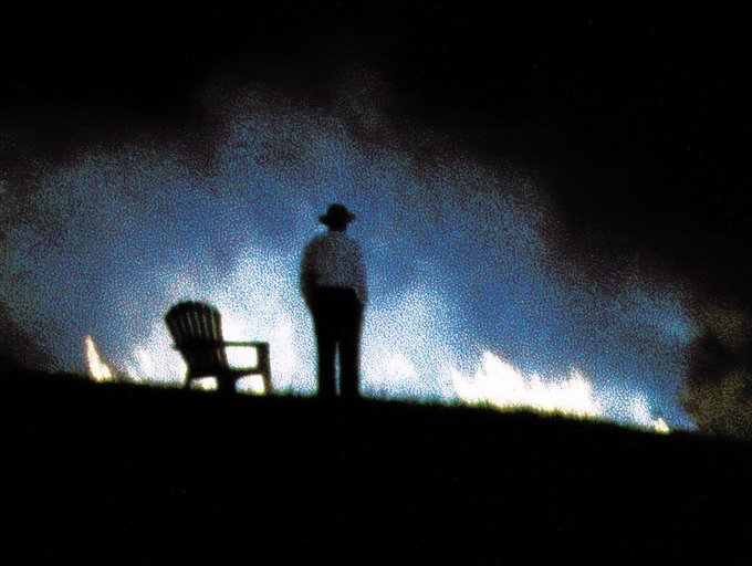 A silhouette of a person wearing a hat standing next to an empty plastic chair at night, illuminated by flames and smoke in the background.