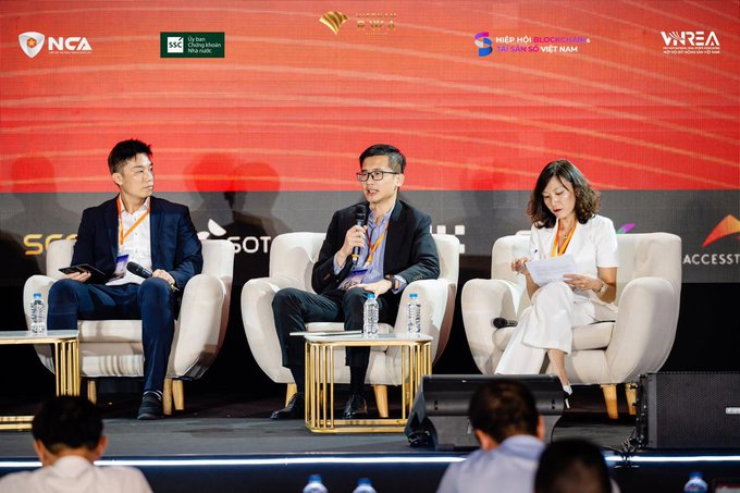 Three panelists sit on stage in white chairs during a conference, with a red backdrop displaying logos including NCA, UK Aid, and 'HIỆP HỘI BLOCKCHAIN TÀI CHÍNH SỐ VIỆT NAM'.