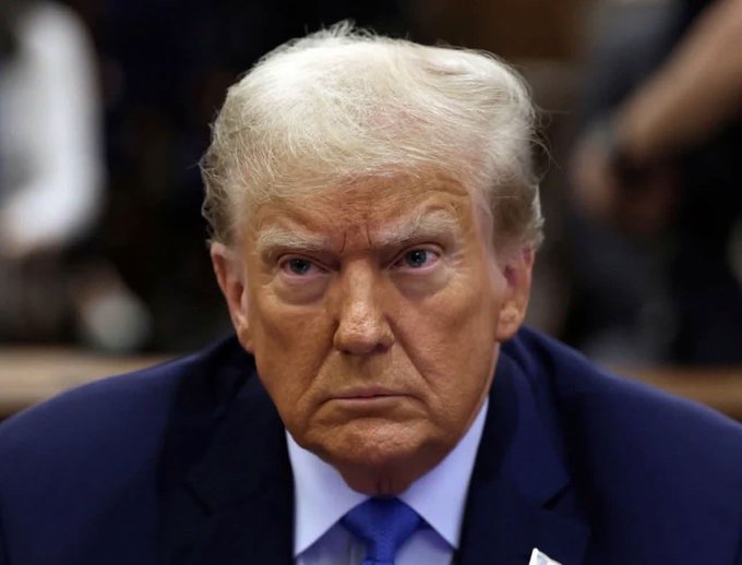 The image shows a man wearing a blue suit, which suggests he might be in a formal setting or an event taking place at a government building such as the State Department. The man has blonde hair styled in an unconventional way with bangs covering his eyes. In the background of the photo, there are blurred figures that could potentially represent other individuals present in the scene but not visible enough to discern their actions. There is also text in the image that appears to be a description or commentary about Donald Trump's upcoming speech at the State Department.