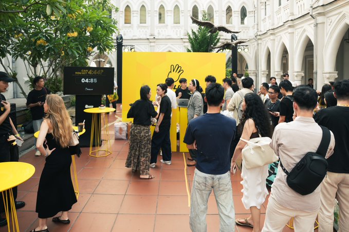 A crowd of people gathers at an outdoor event with white Gothic-style arched architecture, centered around a bright yellow installation and a countdown timer displaying '04:05'.