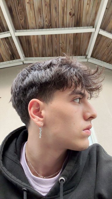 A side profile of a young man with curly dark hair, wearing a silver cross earring and a black hoodie.