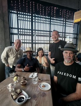 Group photo of five people gathered around a restaurant table with food and drinks; one man on the right wears a black shirt reading 'FREEDOM' and a hat.