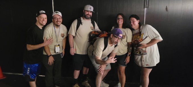 A group of five people posing together in matching beige t-shirts at what appears to be a convention or event, with one person bending over and making a hand gesture.