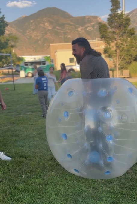 Photograph of a man inside a large clear inflatable bubble ball on a grassy park, with children, a food truck, and mountains visible in the background.