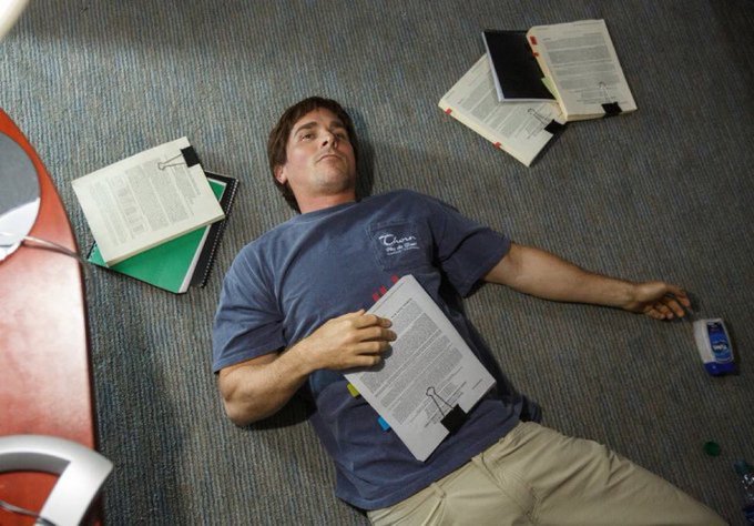 A man lies on a carpeted floor scattered with books and papers, clutching a document and appearing asleep or exhausted in what looks like a study area.
