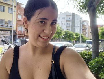 A selfie of a smiling young woman with dark hair wearing a black tank top, taken on a street with buildings and parked cars behind her.