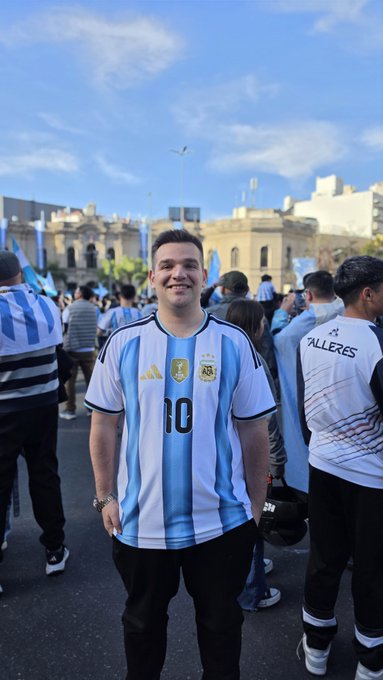 A smiling man wearing an Argentina national football team jersey (number 10) stands in a crowd of fans, likely at a World Cup celebration or match event.