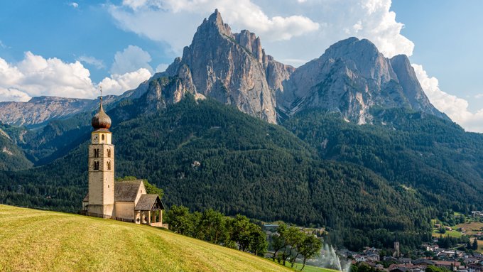 A small church with a tall bell tower sits on a grassy hill overlooking the Dolomite mountains, a tiny settlement nestled in the valley below. The image evokes a sense of timeless serenity and immovable grandeur, humorously mirroring the crypto post's suggestion that Bitcoin would remain stuck in a stagnant $200 range forever, as if frozen in time like the ancient alpine landscape.