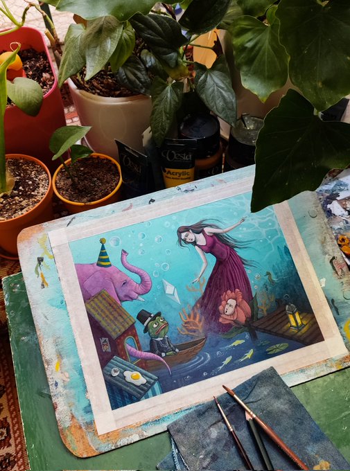 The image features an art project with a painting of a woman dancing underwater, surrounded by various objects such as boats, elephants, fish, and other sea creatures. The painting has been completed and is ready for display or sale. There are two potted plants in the scene - one on the left side and another on the right side. A cup can be seen near the center of the image, possibly containing paint or water to clean brushes. Additionally, there's a book placed towards the bottom-left corner of the painting. The art project is displayed on a green table with a blue mat underneath it.