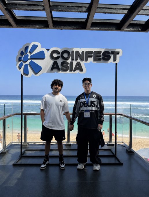 Two young men pose for a photo in front of a 'COINFEST ASIA' sign with a beach and ocean visible in the background.