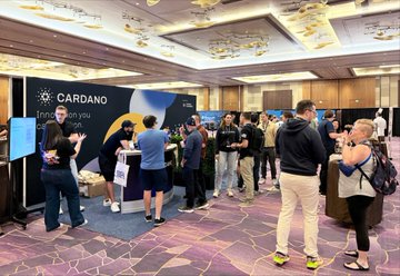 A photograph of a crowded event space where attendees are gathered around booths, including one prominently labeled 'CARDANO Treat for you'.