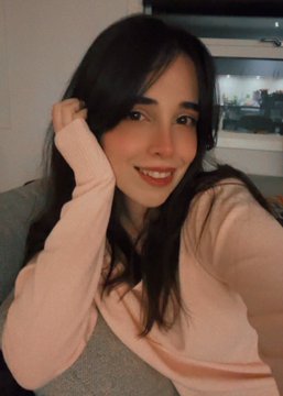 A selfie of a smiling woman with long dark hair wearing a pink and grey top, posing indoors.