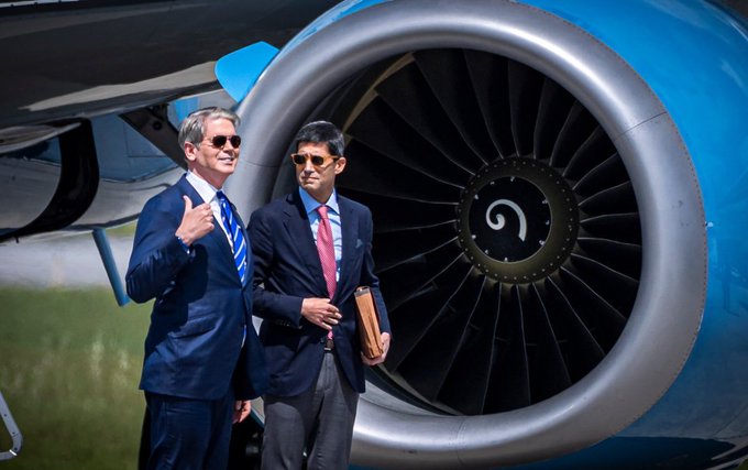 Two men in business suits and sunglasses stand in front of a large blue jet engine, one giving a thumbs-up gesture while holding a folder.