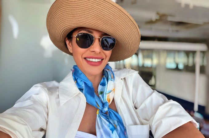 A selfie of a smiling woman wearing a tan sun hat, aviator sunglasses, white blouse, and a blue floral silk scarf, taken inside what appears to be a vehicle.
