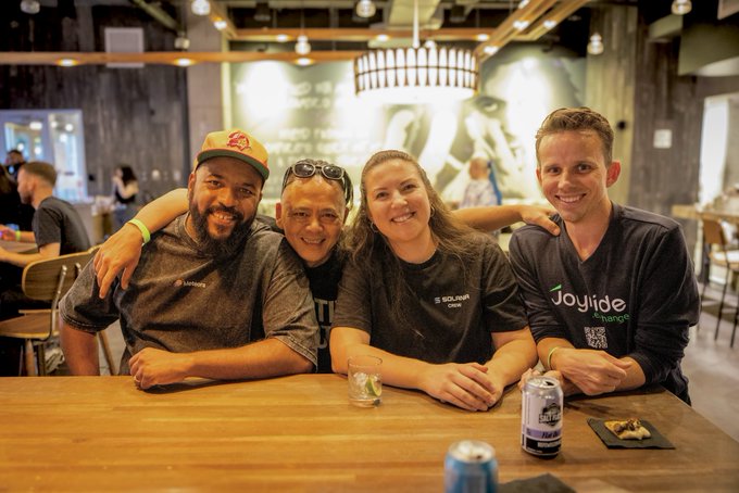 Four Solana team members—two wearing "SQUAD Ops" shirts—pose together at a wooden table with drinks, smiling for the camera in what appears to be a casual gathering space. The image captures the community-driven culture mentioned in the post about SLC and LA events strengthening Solana's community bonds.