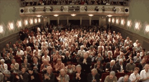 The image shows a large crowd of people sitting in rows of chairs at an event or gathering. Many individuals are clapping their hands together, indicating that they are engaged and enjoying themselves. The audience appears to be seated in front of a stage where a speaker is giving a presentation. The overall atmosphere seems lively and energetic as the attendees participate actively in the event.
