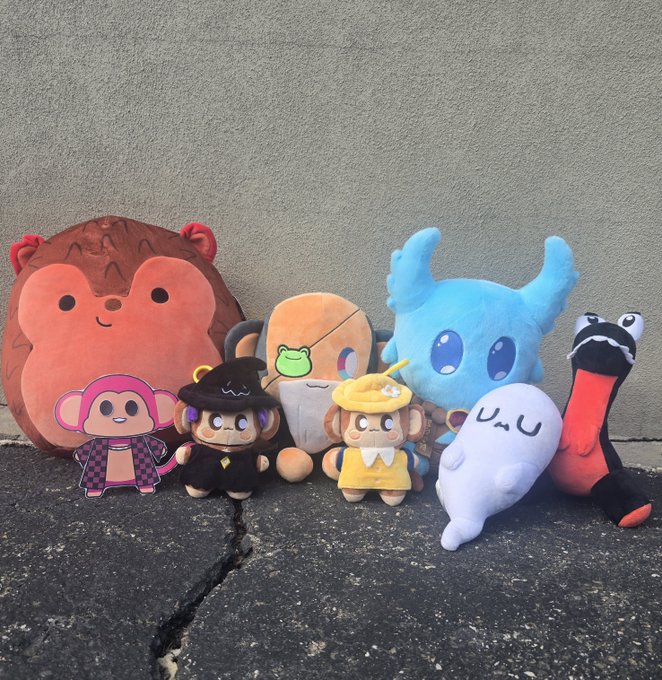A group of colorful plush toys and character pillows is arranged against a gray wall on dark pavement.