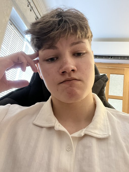 A close-up selfie of a young person with short brown hair wearing a white collared shirt, resting their hand near their head with a neutral expression.
