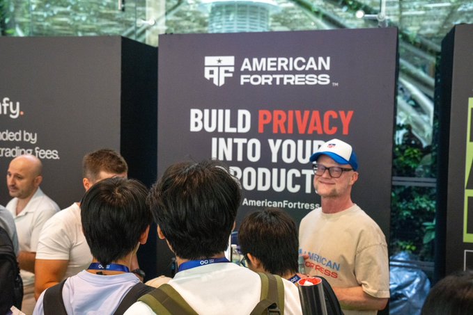 A man wearing a white and blue American Fortress cap and t-shirt stands at a trade show booth with a large sign reading 'BUILD PRIVACY INTO YOUR PRODUCT' and 'AmericanFortress'.