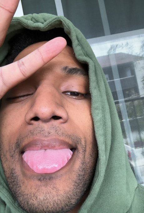 A young man in an olive-green hoodie poses for a close-up selfie, winking with one eye, holding two fingers to his forehead, and sticking out his tongue in a silly, playful expression. The caption references the crypto influencer ANSEM ($ANSEM), matching the meme-like, irreverent selfie style often seen in his community posts.
