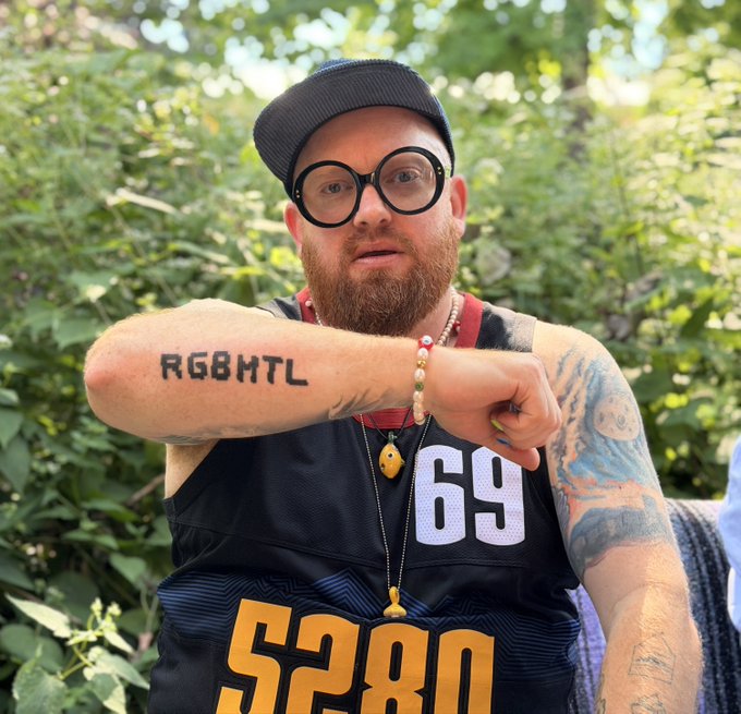 A bearded man wearing round glasses, a cap, and a black jersey with the number 69 shows off a forearm tattoo reading 'RG8HTL' while standing in front of greenery.