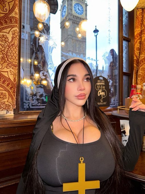 A young woman with long dark hair wearing a black hooded top and a large gold cross necklace poses indoors, with a clock tower visible through the window behind her.