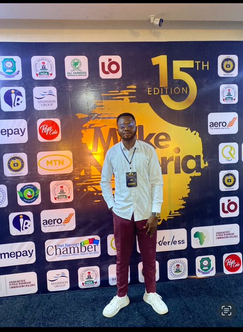 A man wearing glasses, a white shirt, and maroon pants poses in front of a "15TH EDITION" backdrop covered with sponsor logos including MTN, PiggyVest, and Aero for what appears to be a "Mike Oria" event.