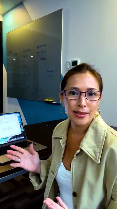A woman with glasses, wearing a beige jacket, gestures while speaking in front of a whiteboard with writing on it and an open laptop on the desk beside her.