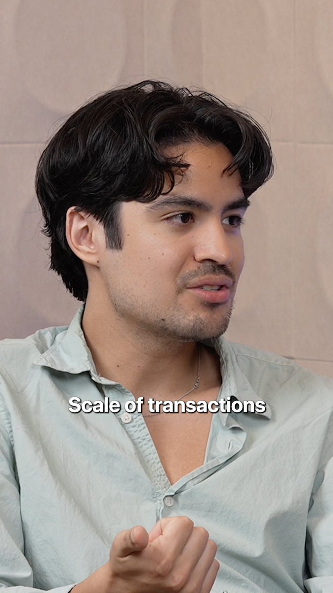 A young man with dark wavy hair and light stubble, wearing a pale blue button-up shirt, speaks to someone off-camera while gesturing with his hands. The text "Scale of transactions" appears across the image, tying into the post's discussion of blockchain's ability to solve transactional bottlenecks in large-scale marketplaces.