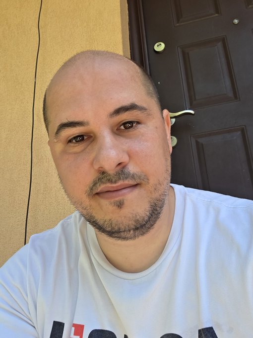 A man takes a casual selfie in front of a dark wooden door, wearing a white t-shirt that appears to have partial "I ❤️" lettering visible at the bottom. His relaxed morning look fits perfectly with the "GM" (Good Morning) greeting, and his slightly sun-touched appearance hints at wanting to get back into vacation mode.