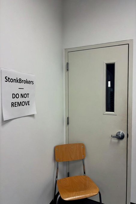 A simple room corner with a wooden chair in front of a white door, with a paper sign on the wall reading 'StonkBrokers — DO NOT REMOVE'.