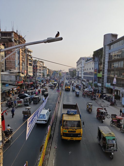 The image shows a busy city street filled with various vehicles such as cars, trucks, and motorcycles. There are also people walking along the sidewalks near the buildings. A bird can be seen perched on top of one of the buildings. The scene captures the hustle and bustle of urban life in an Indian city.