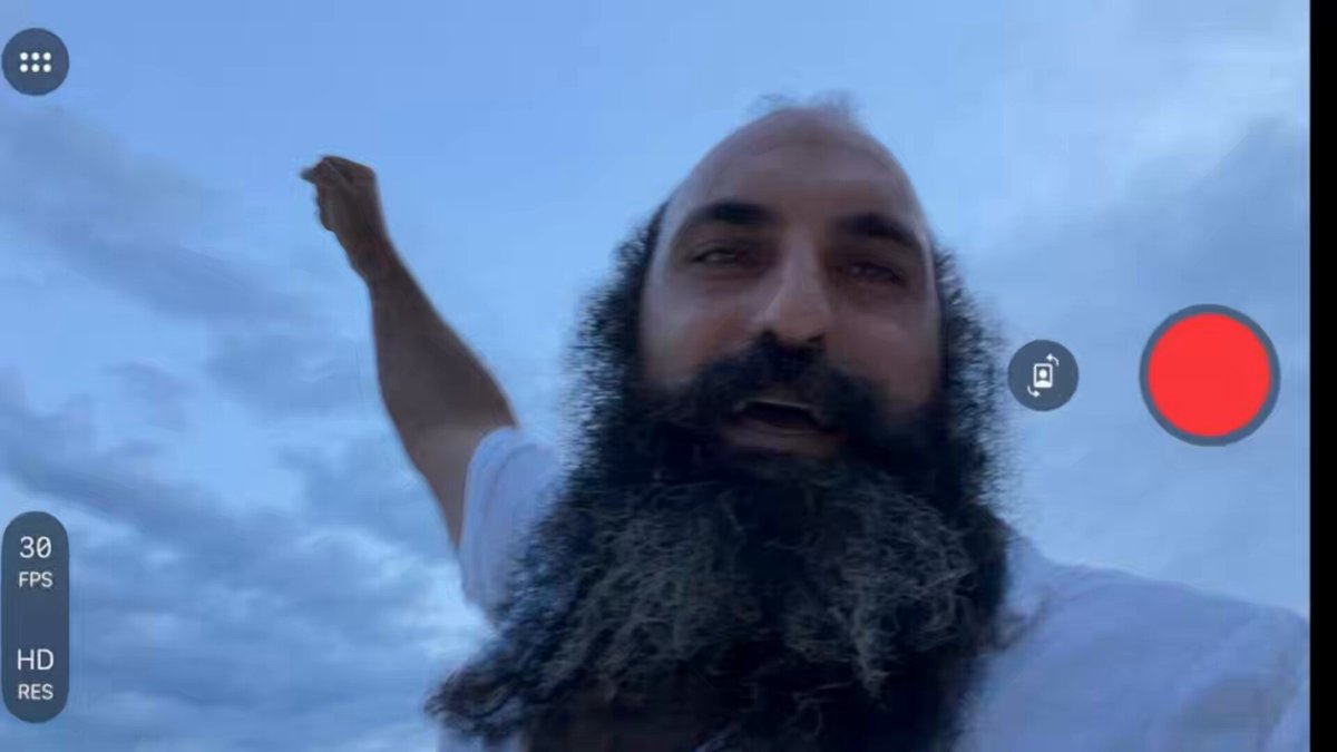 The image shows a man with a beard wearing a white shirt, holding up his arm as if he's waving or making a gesture. The background of the photo features a cloudy sky, which contrasts with the man's facial hair. There are two red circles in the bottom right corner of the image, and they appear to be related to Bitcoin.