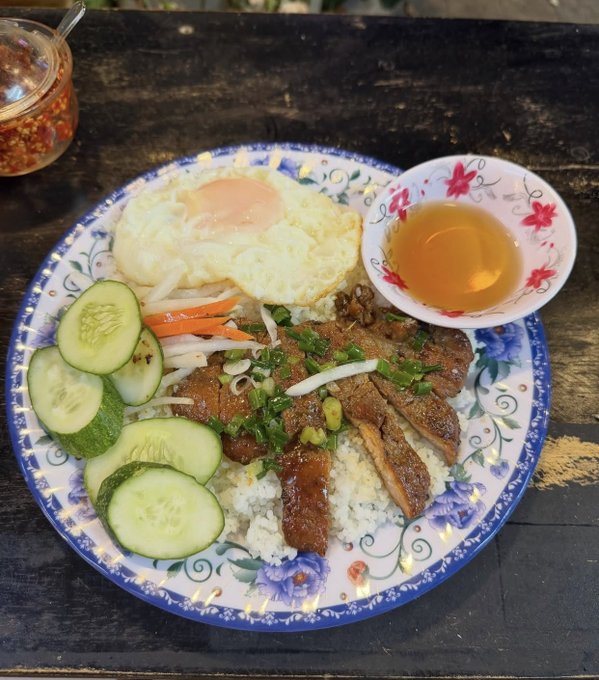 A plate of Vietnamese cơm tấm featuring grilled pork, a fried egg, cucumber slices, pickled carrots, and a side of fish sauce on a decorative blue floral plate.