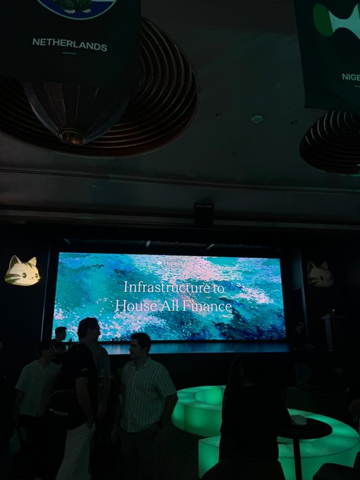 A dimly lit event space features a prominent screen displaying 'Infrastructure to House All Finance', with attendees mingling below hanging wooden ceiling decorations.