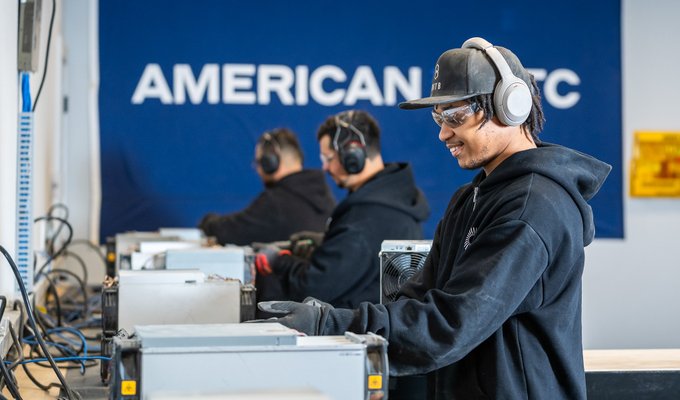 A smiling young man wearing safety glasses, headphones, and a black hoodie working on computer hardware, with two other workers in the background and a blue 'AMERICAN' banner on the wall.