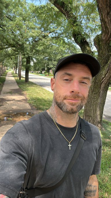 A man wearing a black cap, dark t-shirt, and cross necklace takes a selfie on a tree-lined sidewalk.