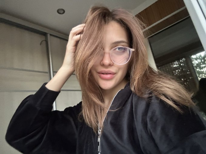 A young woman with long brown hair, pink-tinted glasses, and a black jacket takes a selfie while adjusting her hair, visually embodying her tweet "Elon, you're gonna love this: I'm a girl on X" as she playfully introduces herself to Elon Musk on the platform.